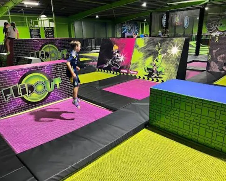 A wide shot of the inside of Flip Out that features many colourful neon trampolines built into the ground, with children jumping on them and some bright green obstacles and standing blocks amongst the trampolines.