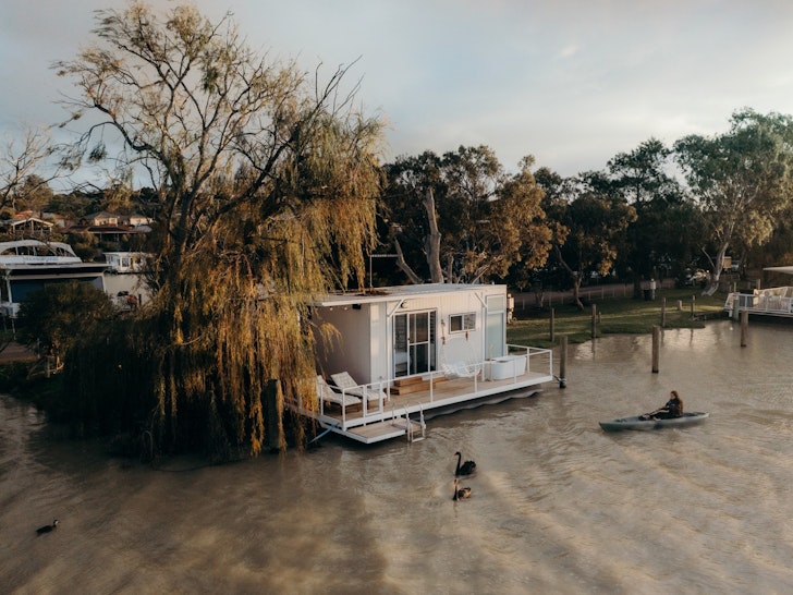 A small white tiny home floats on a river while a person kayaks in front of it.