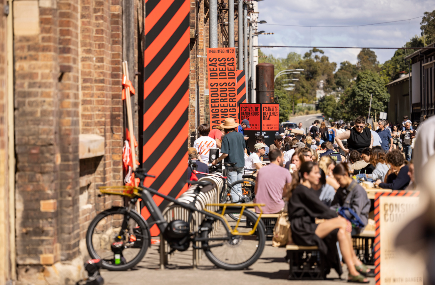 Festivalgoers gather outside Carriageworks during the Festival of Dangerous Ideas, with crowds dining, cycling and exploring the precinct beneath the festival's signature orange-and-black branding.