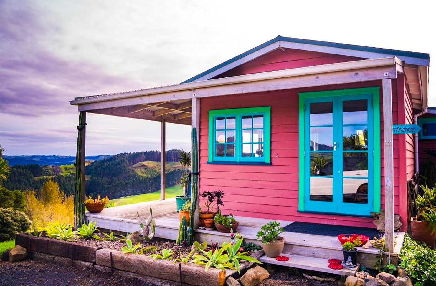 A pink cabin with peppermint and baby blue detailing looks out on rolling hills at Fridas Accommodation, one of the best places to stay in Raglan. 