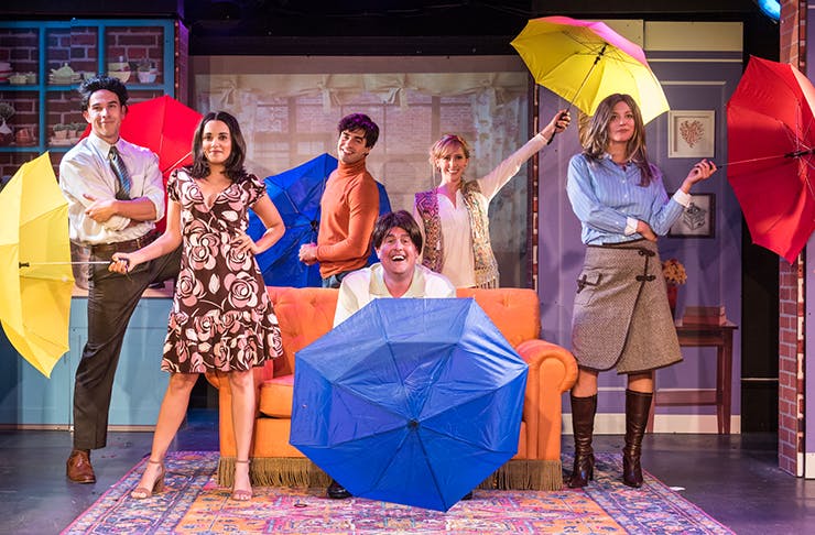 Three actors dressed as the characters from Friends, holding umbrellas and standing on a stage.