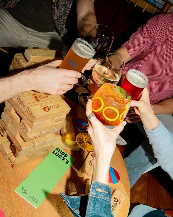 People at Miss Lucy's clink cocktails over a small round wooden table covered in board games.