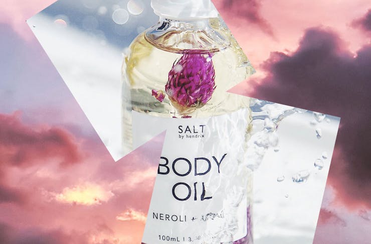 A collage of two images, the background is pink clouds, and the front is a bottle of body oil. 