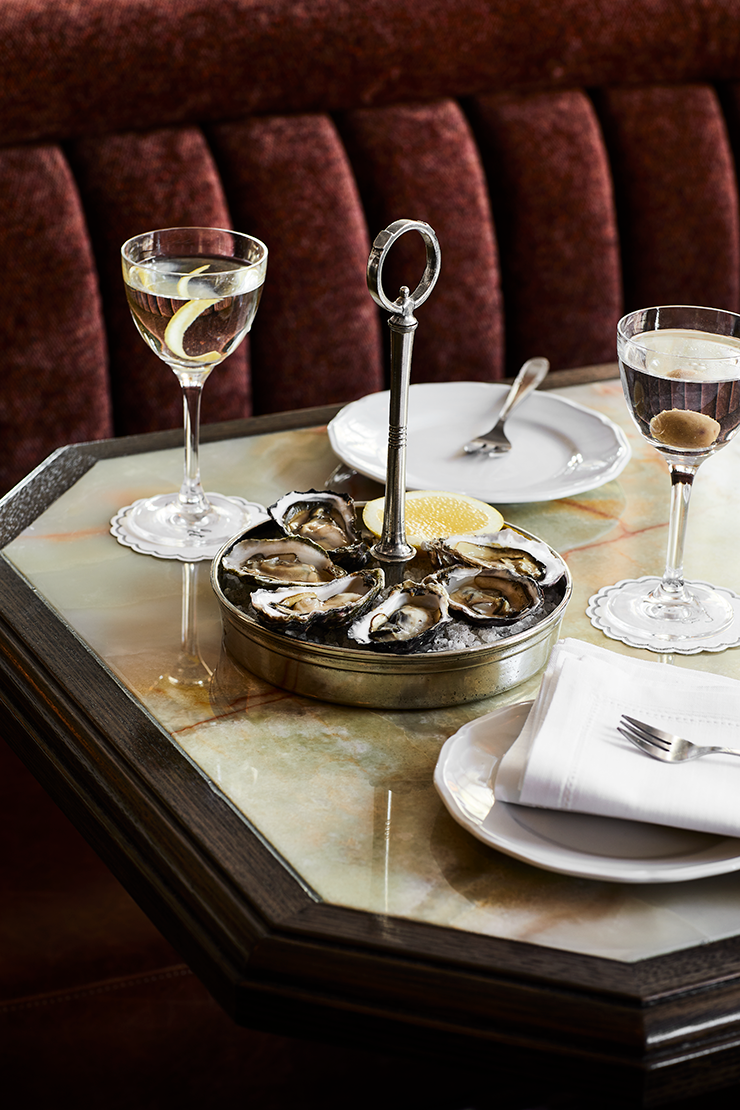 A silver plate holding oysters and surrounded by cocktails garnished with citrus peel.