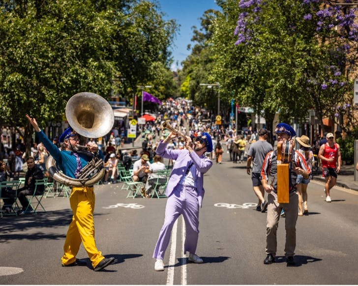 On an outdoor street full of people, the image focuses on three musical performers dressed in colourful outfits and in the middle of playing music on their instruments.