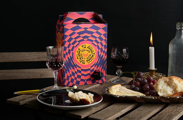 A colourful box of cask wine on a table next to a plate of cheese and ornate wine glasses. 