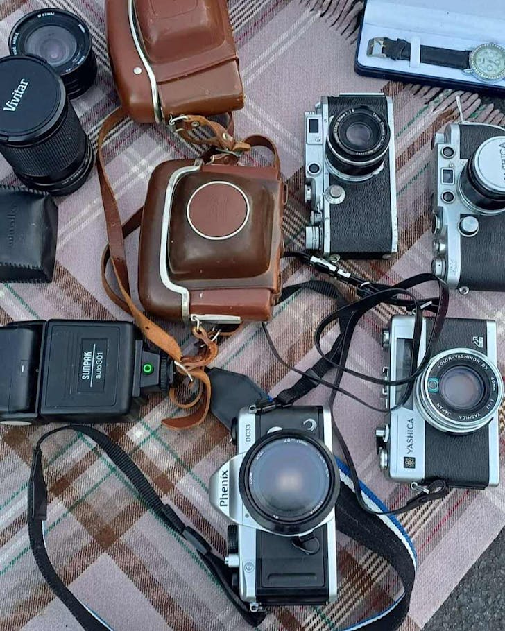 Old film cameras and accessories lay on a table covered in a flannel blanket, waiting to be sold.