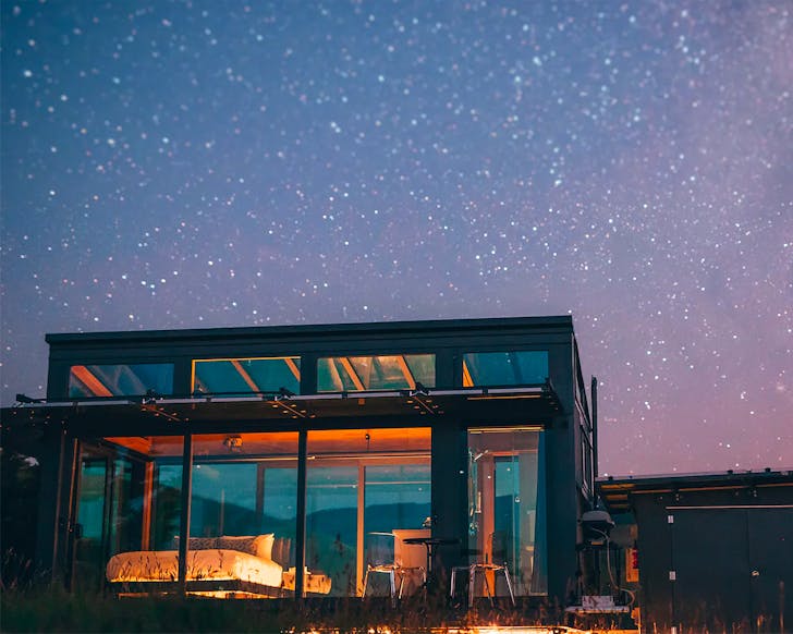 A glass cabin sits on a hill at night time, lit from within. Overhead a billion stars shine brightly. 