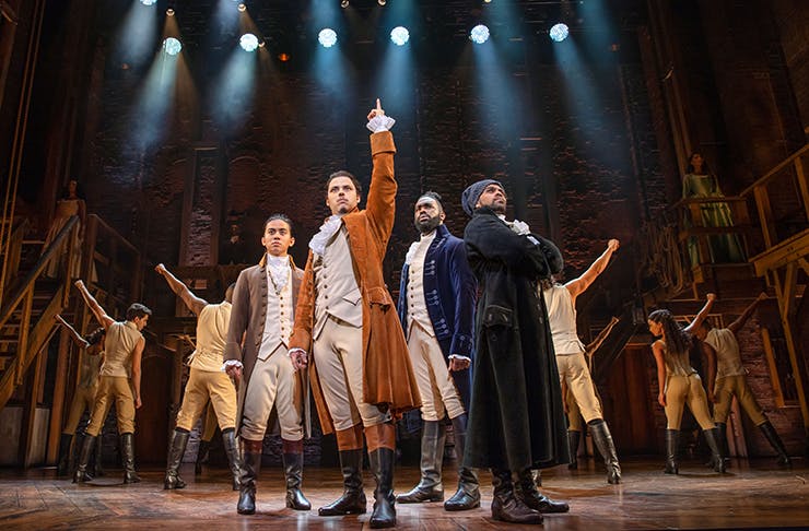 The cast of Hamilton on a stage under lights.