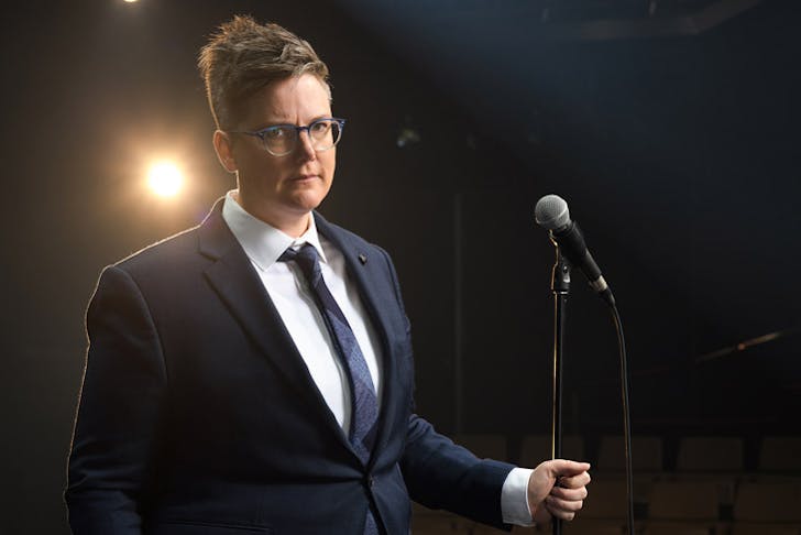 A portrait of Australian comedian Hannah Gadsby on stage wearing a blue suit. 