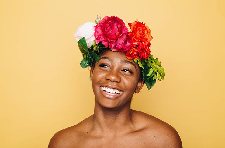 A person standing in front of an orange wall with a big smile on their face and flowers in their hair.