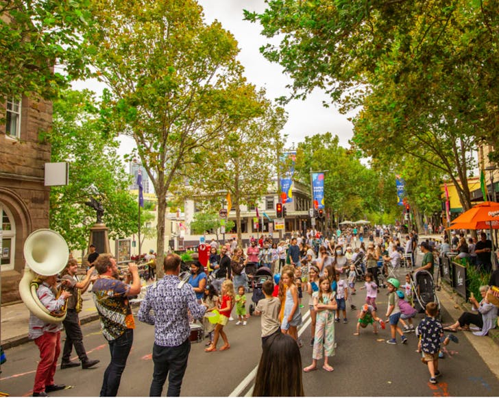 A wide shot showing some distance down an outdoor street. The image shows children dancing and watching the live musicians in the forefront who play hand held drums and wind instruments.