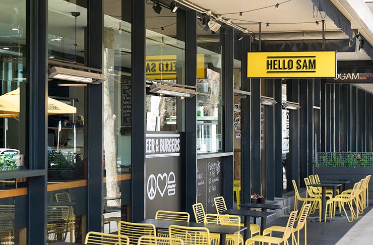 The exterior of a restaurant with floor to ceiling windows and yellow seats out the front. A yellow sign with black text reads 