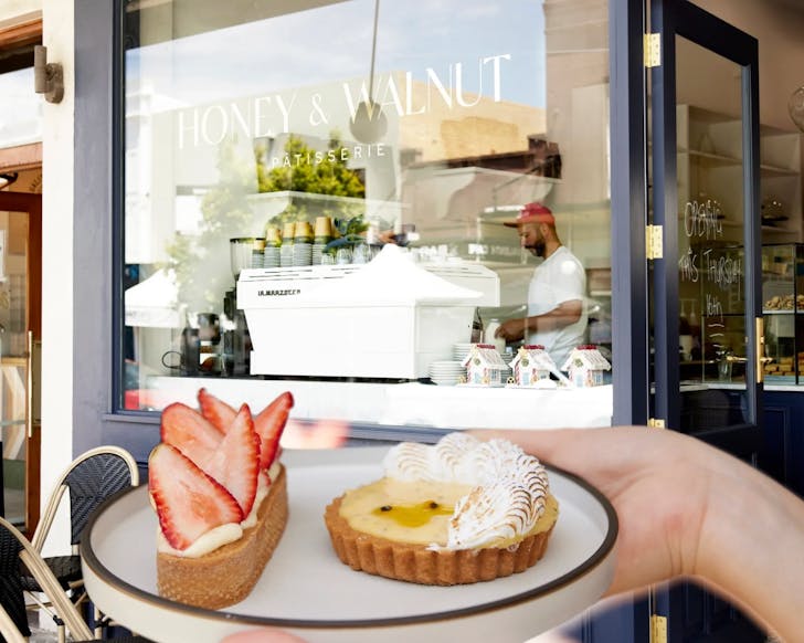 A close up image of a hand holding a plate with two patisserie sweets on it. In the background is the front window of Honey & Walnut Patisserie, where a barista can be seen making a coffee.