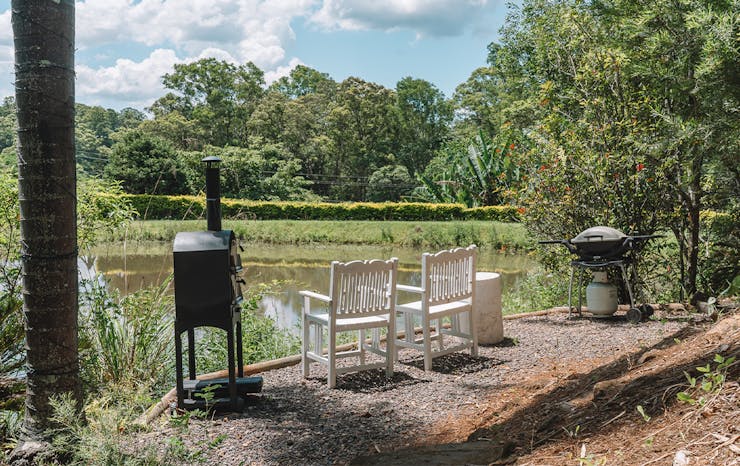 a pizza oven and bbq next to two white chairs overlooking a small dam