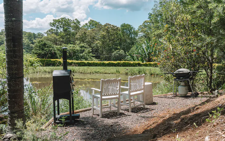 a pizza oven and bbq next to two white chairs overlooking a small dam