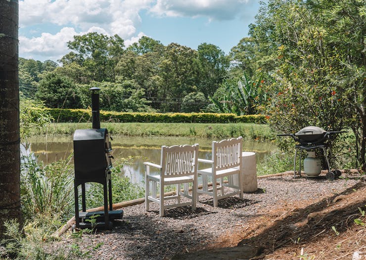 a pizza oven and bbq next to two white chairs overlooking a small dam