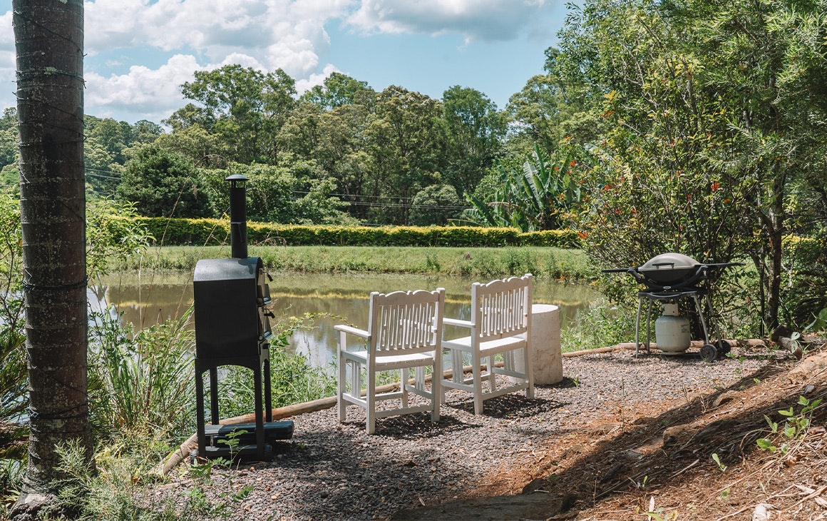a pizza oven and bbq next to two white chairs overlooking a small dam