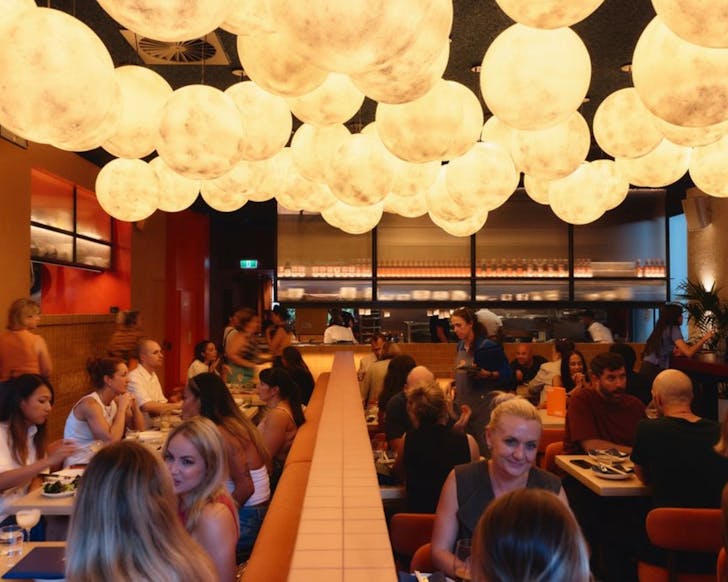 A wide shot of inside the Island Radio venue, with rows of elegant dining tables seated under artsy lights hanging from the ceiling above. Many people sit at the tables eating and talking, while others walk around the space happily.