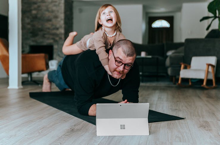 A man tries to hold a plank in front of a laptop while his child hangs onto his neck.