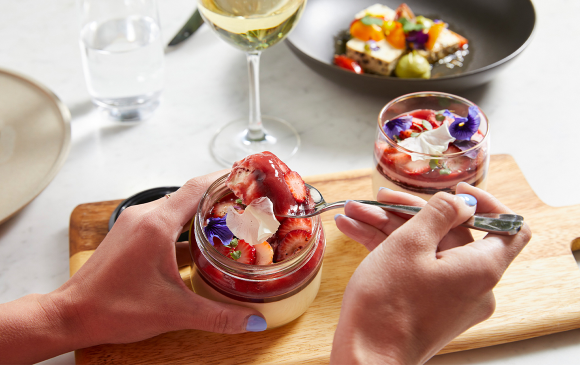 a spoon being scooped into a glass jar of a creamy, strawberry topped dessert
