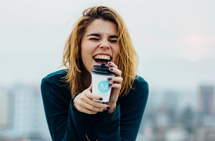 A woman laughing drinking a coffee out of a disposable cup.