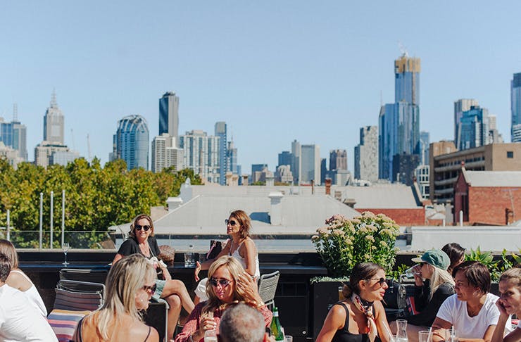 People enjoying a drink on a rooftop bar. Melbourne skyline is in the background.