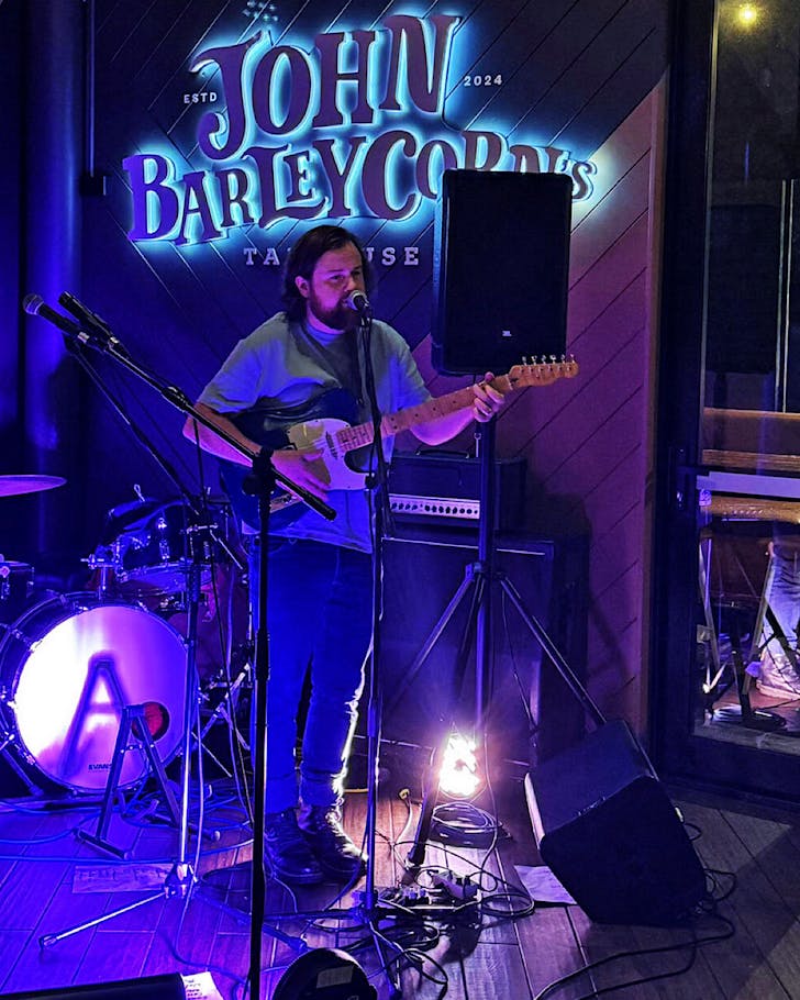someone plays guitar in front of a blue-lit stage and a large 'John Barleycorn's' sign at a live music night.