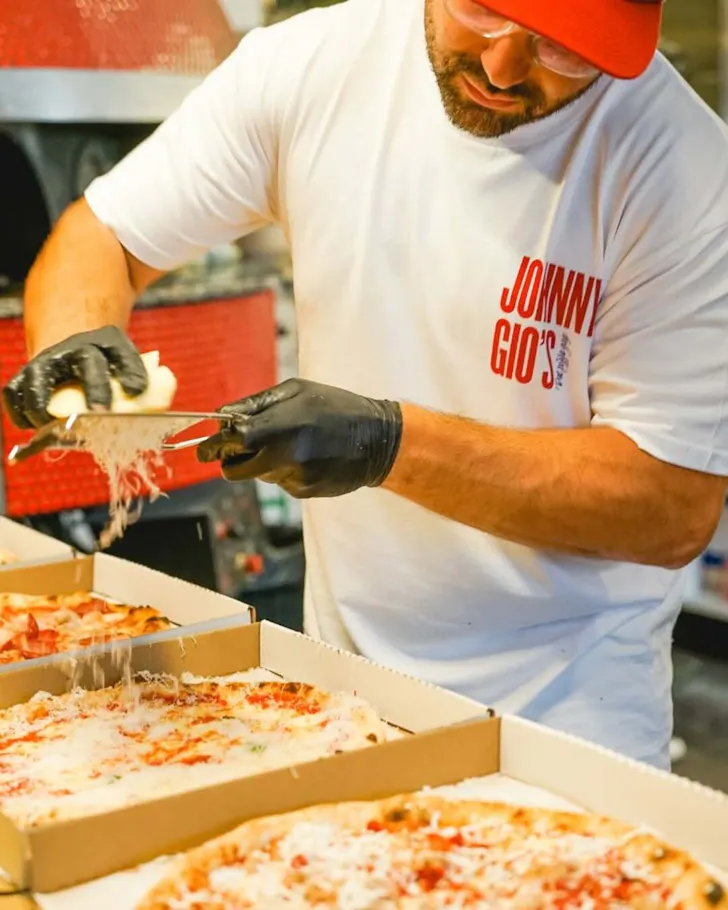 A close up shot of a man's torso as he grates cheese on top of fresh pizza. The pizza are sitting in boxes at the bottom of the image, with different toppings including red sauce, meats, and cheese.