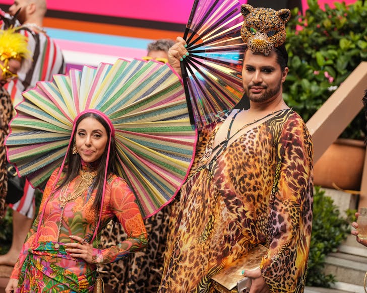 Two people dressed in vibrant, animal-print kaftans and bold accessories at a poolside Mardi Gras event. One wears a colourful, fan-shaped headdress and gold jewellery, while the other sports a sequined leopard headpiece and iridescent fan. The