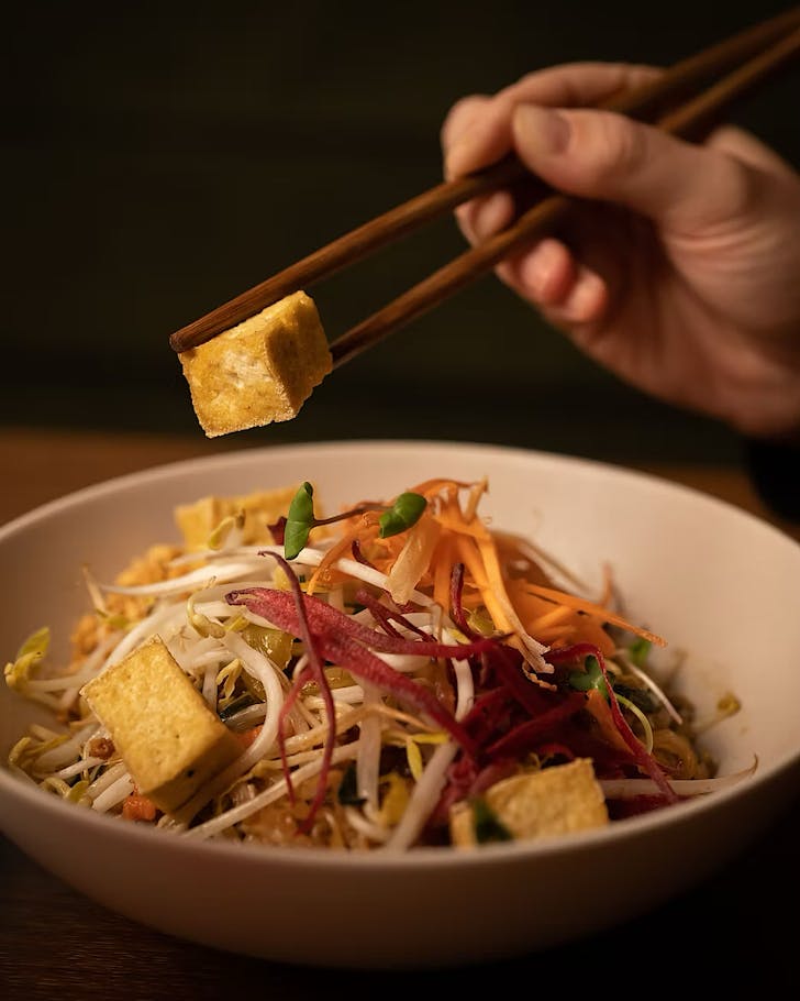 Someone uses chopsticks to pick up a cube of tofu from a bowl of Pad Thai.