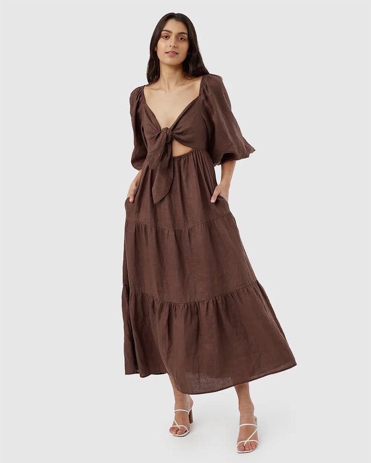 A woman wears a dark brown full length dress with ample sleeves and two ties that create a bow allusion over her bust. 