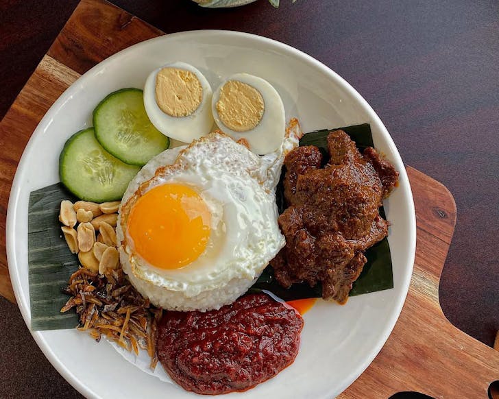 A plate of rice, a fried egg, a halved boiled egg, slices of cucumber, anchovies, beef rendang and sambal.