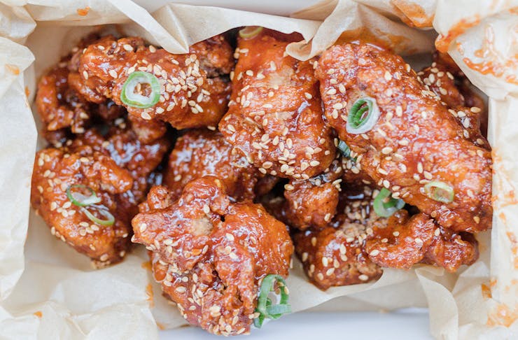 Saucy, sticky boneless fried chicken in a takeaway box, sprinkled with sesame seeds and shallots.