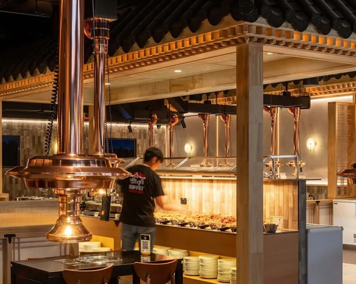 A wide shot of a section of the Korean BBQ restaurant, where the focus is on a man stands in the middleground who stands at a window of display all-you-can-eat food, his back to the audience. It is night and the lighting is warm, with some BBQ tables shown in the foreground.