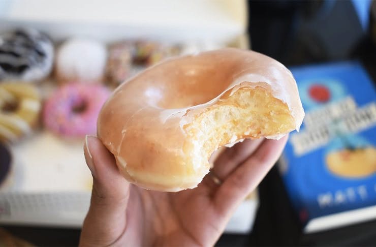 A hand holds a glazed doughnut with a bite taken out of it.