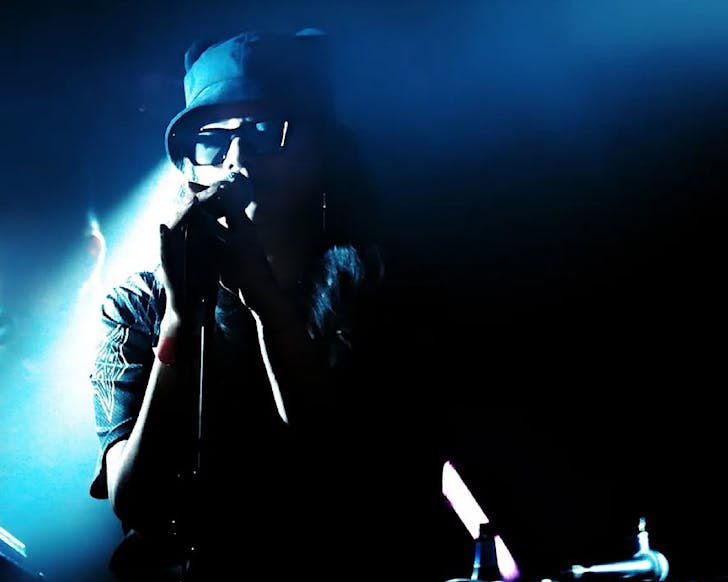 Ladi6 performs on a dark, blue-lit stage in a bucket hat and sunglasses.