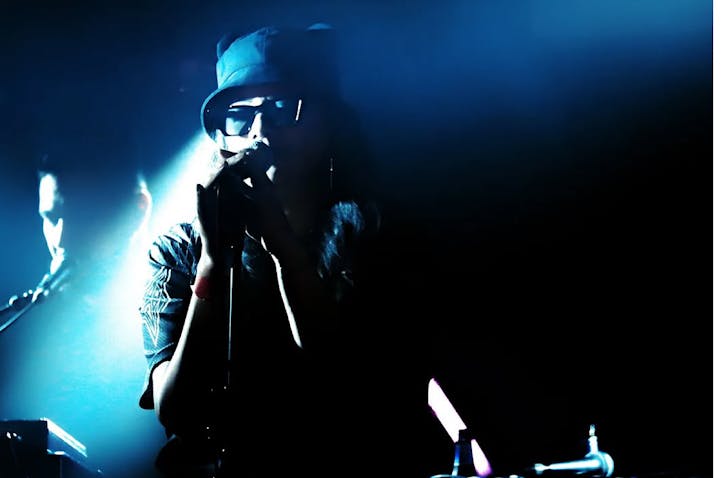 Ladi6 performs on a dark, blue-lit stage in a bucket hat and sunglasses.