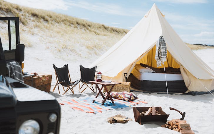 A bell tent with bed visible inside, fire pit, chairs and the side of a cool looking truck.