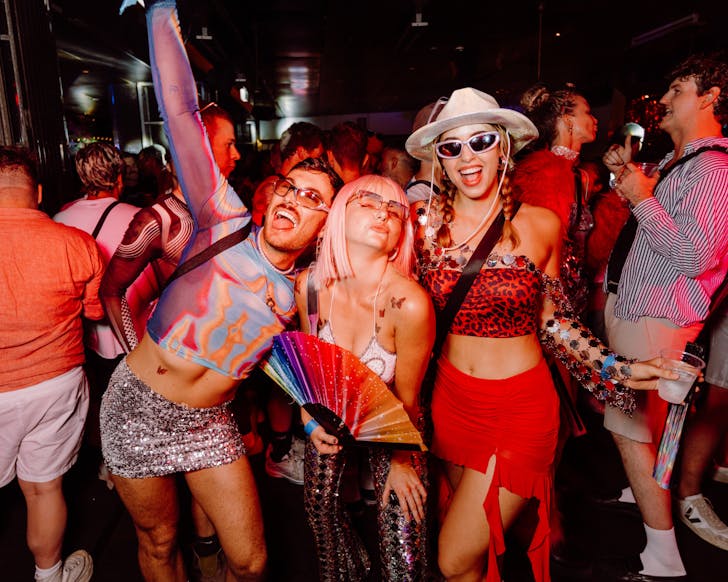 A lively crowd dances at Sydney Mardi Gras Laneway Party, with three friends in vibrant outfitsâsequins, leopard print, and a rainbow fanâposing joyfully under red lights at The Beresford.