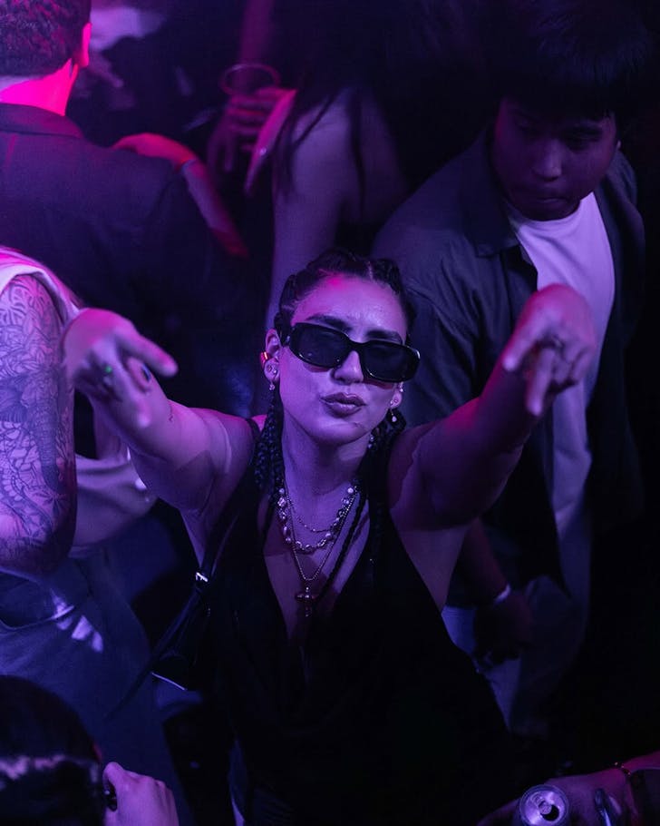 Someone at a Latin XL gig poses for the camera in dark sunglasses on a pink-lighted dance floor.