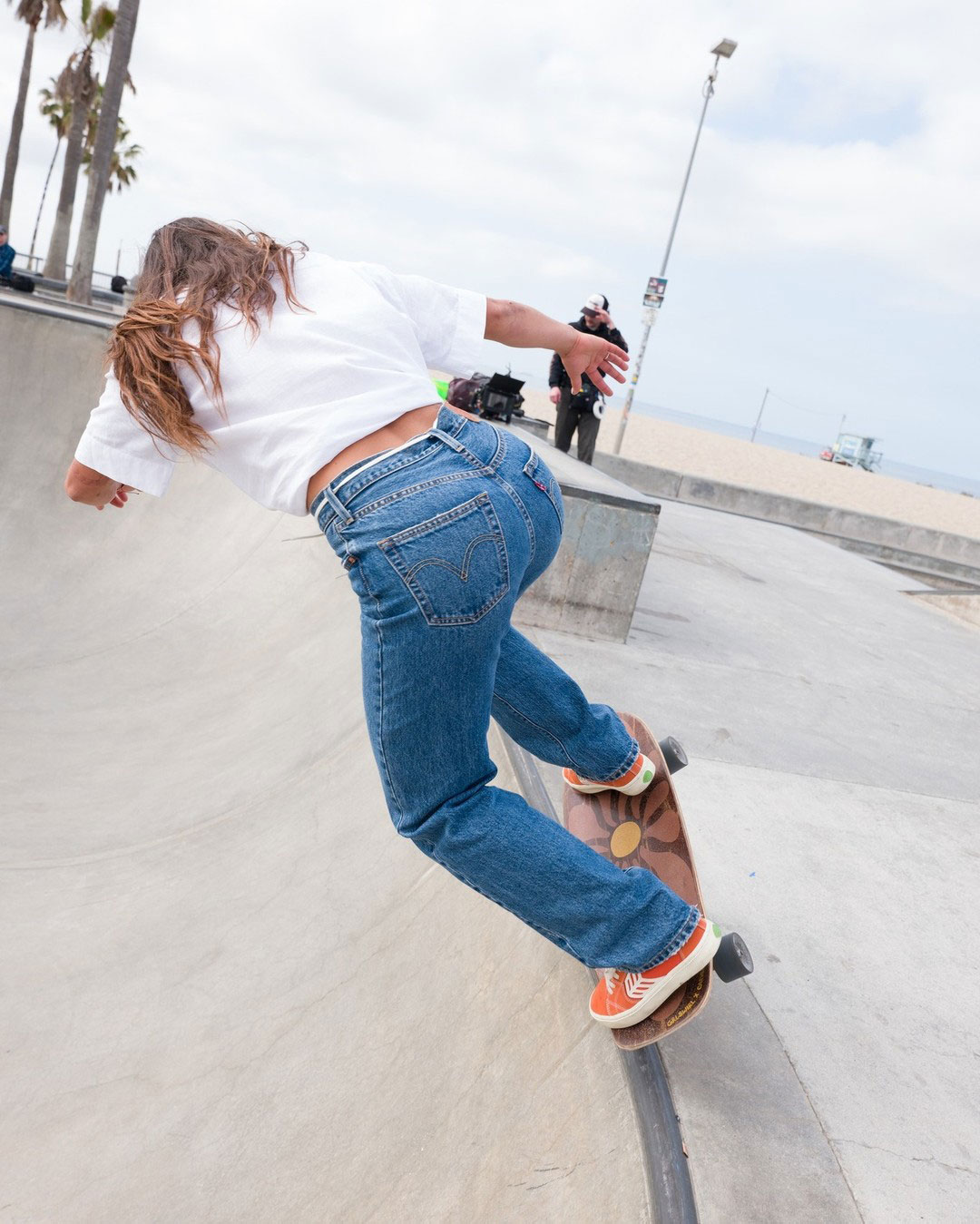 A female skating on the rim of a skate bowl wearing a white t-shirt and blue Levi's jeans.
