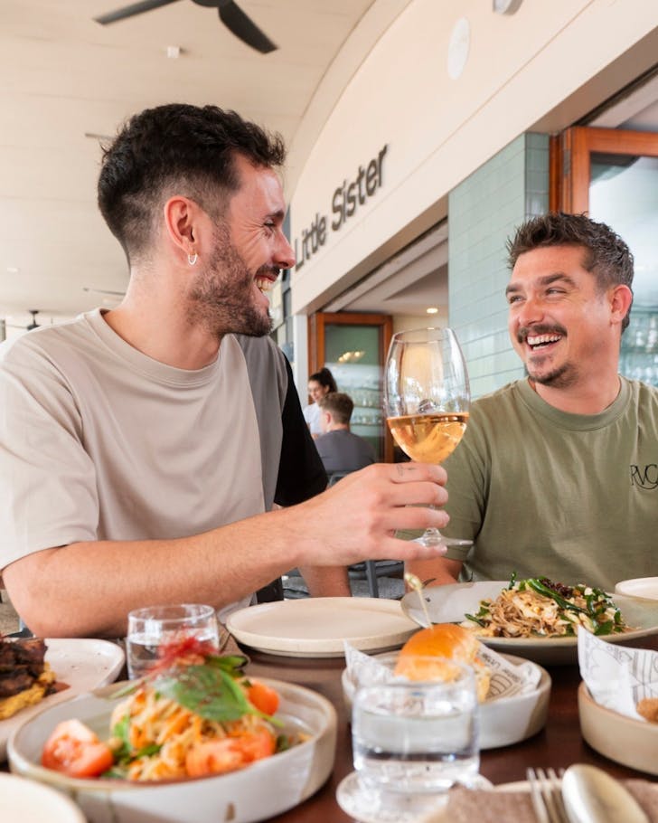 Two friends clink wine glasses over a table of Asian street food at Little Sister cafe in Cairns.
