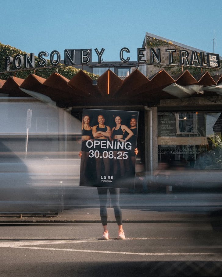 An LSKD employee holds up an opening sign in front of Ponsonby Central, concealing the top half of their body, as traffic whizzes by in a motion blur.