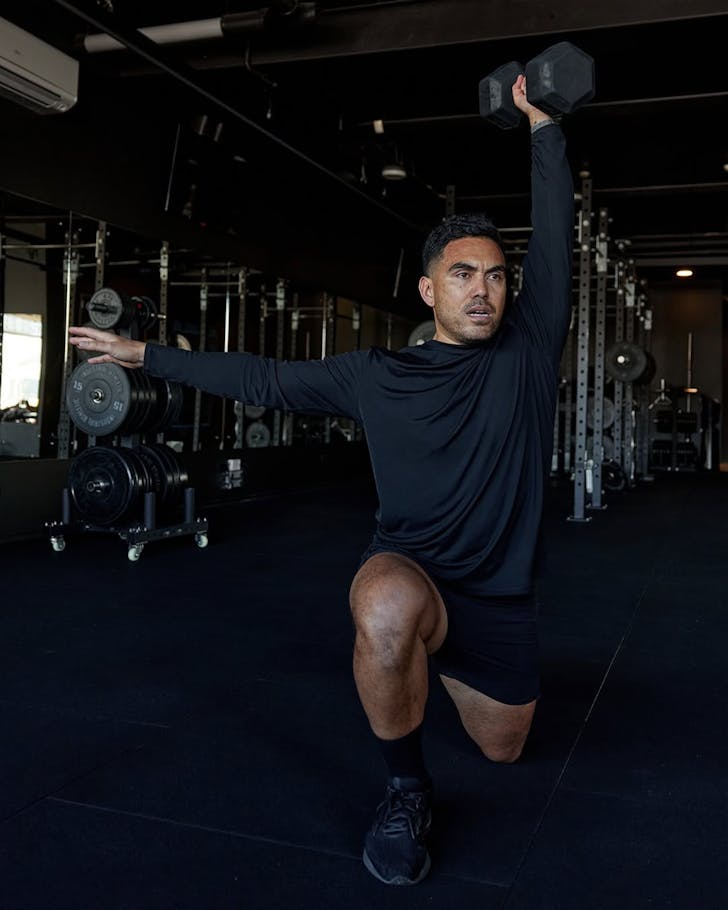 Store athlete Tauvaga Siolo dressed in black LSKD gear does a single let dumbell squat overhead press in a gym.