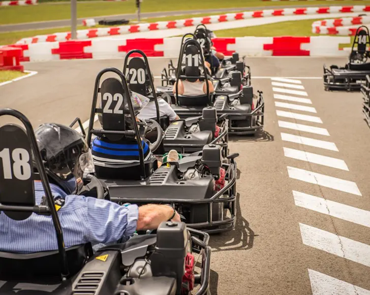 A close up of people in go-karts lined up and ready to race. We see the back of their karts and heads, wearing helmets and there's the beginning of the race track in the foreground and background.