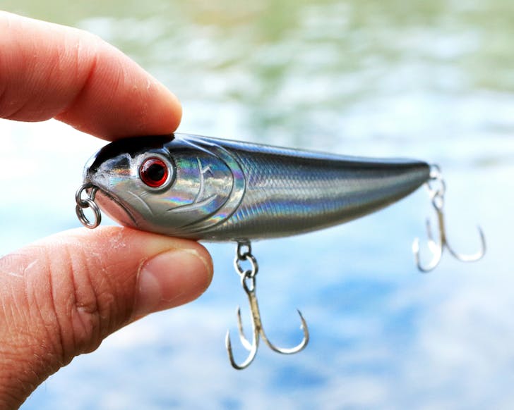 Close-up of a person's hand holding a fishing lure in front of a body of water during the daytime.