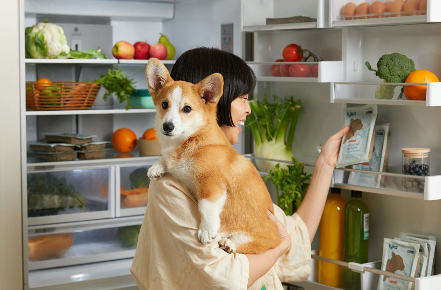 A woman holding her dog opening a fridge to get packets of Lyka fresh food for dogs. 