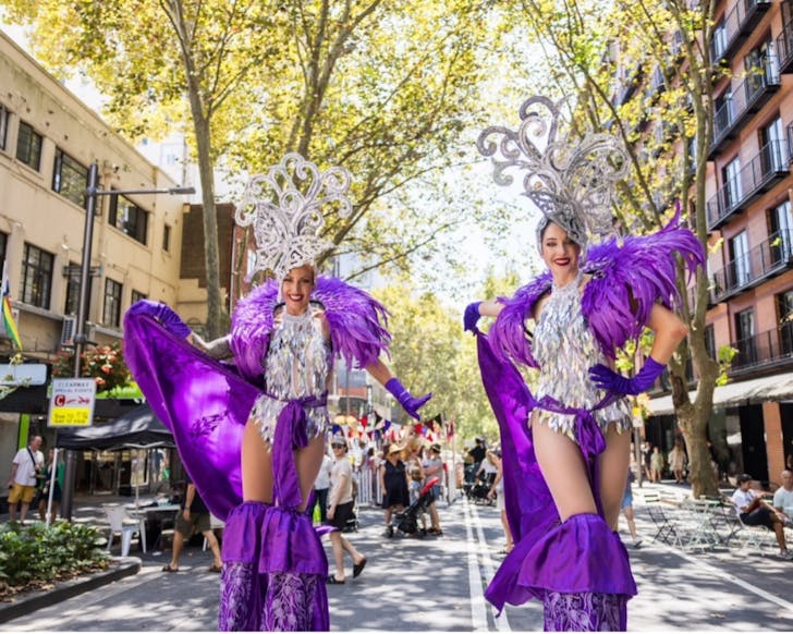 Two women performers are the main focus of the image, dressed in glittery purple and silver costumes and smiling. They are walking down a Sydney street smiling, with the day bright around them.