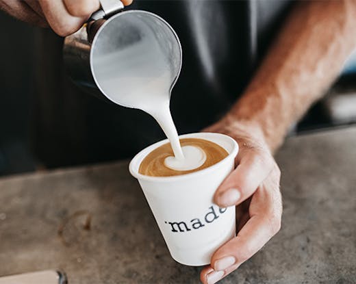 A close-up of two hands holding a white coffee cup that says 'made' on it while pouring frothing milk in the cup.
